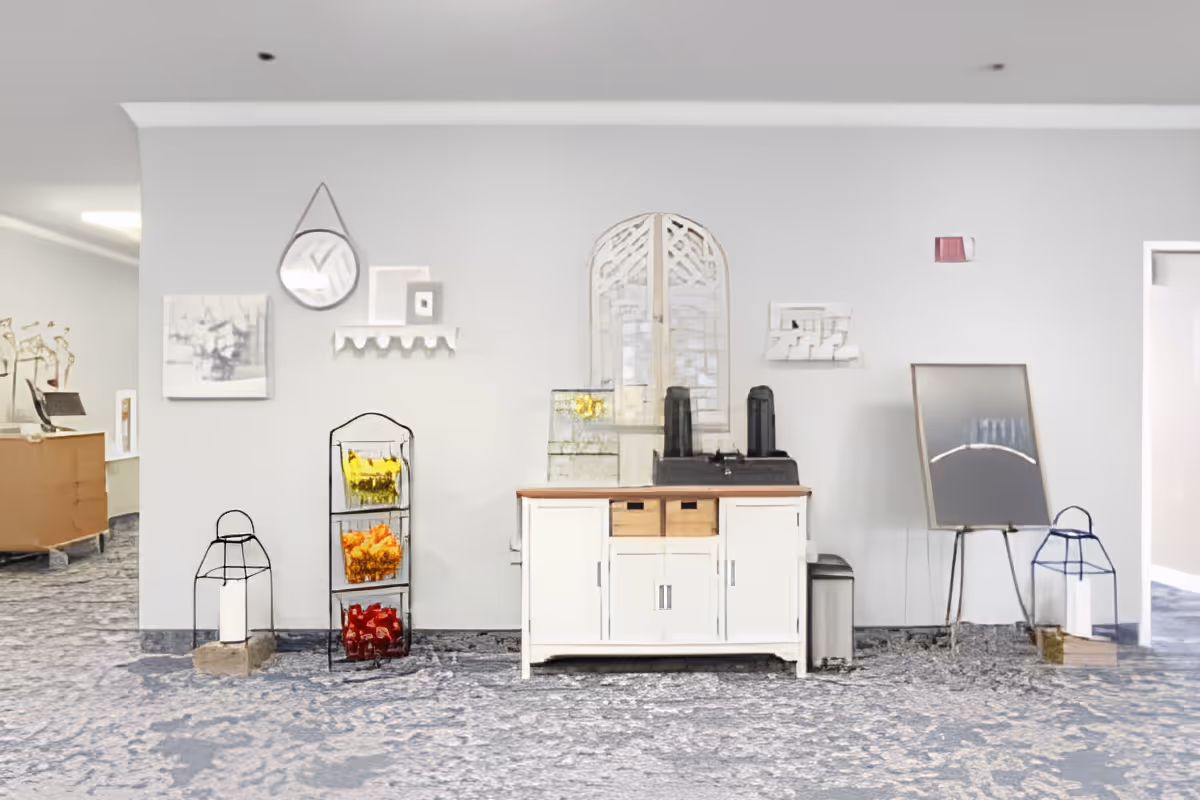 A light gray wall with a white cabinet in the center holding two black coffee dispensers and a glass water dispenser. To the left of the cabinet is a three-tiered rack with yellow, orange, and red decorative items. On the wall above the cabinet are a round mirror, a small shelf, and framed artwork. On the floor to the left and right of the cabinet are large candle holders with white candles. To the right is an easel with a blank canvas or board. The floor is covered with a patterned blue and gray carpet.