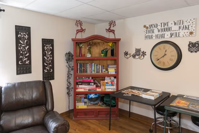 A cozy room corner with a brown leather armchair on the left, a red wooden bookshelf filled with books and board games in the center, and two black tables with puzzles and games on the right. The walls are decorated with two vertical floral metal wall hangings, a large round clock, owl wall decorations, and a sign that reads 'Spend time with those who make your heart smile.' The floor is wooden.