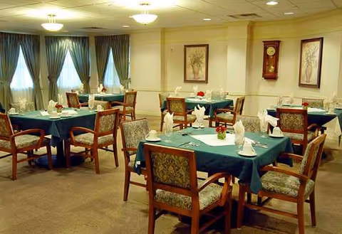 A spacious senior living dining room with teal tablecloths, folded napkins, and wooden chairs.