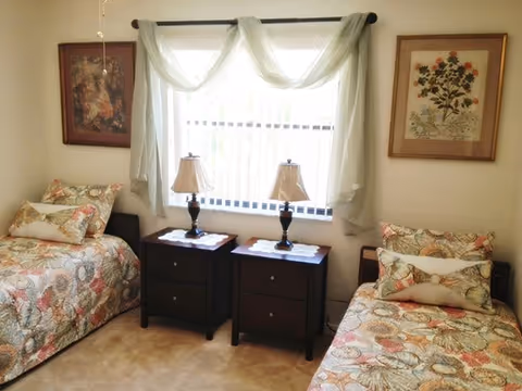 A cozy bedroom with two twin beds featuring floral patterned bedspreads and matching pillows. Between the beds are two dark wooden nightstands, each with a decorative lamp and a doily. A window with sheer white curtains is centered on the wall behind the nightstands. Two framed floral artworks hang on the walls above each bed.