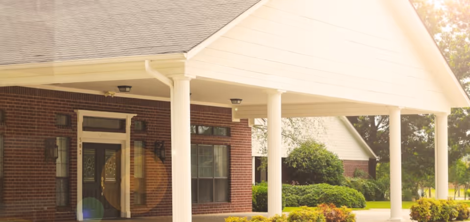 Entrance of a brick building with white pillars supporting a covered driveway, surrounded by green bushes and trees in the background.