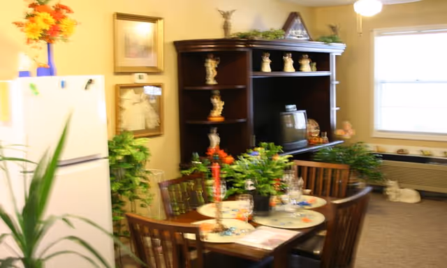 Small interior dining area with a wooden table set for a meal, surrounded by chairs, a dark hutch with decorations and a TV, potted plants, a refrigerator, and a window.