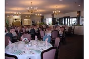 A spacious dining room with round tables set for a meal, each table covered with white tablecloths, pink napkins, and floral centerpieces. The room features chandeliers hanging from the ceiling and large windows allowing natural light to enter.