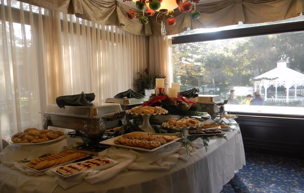 A buffet table set up indoors with various trays of cookies and pastries, covered chafing dishes, and holiday decorations including candles and greenery. Large windows with sheer curtains allow natural light to fill the room, and an outdoor gazebo is visible through the window.