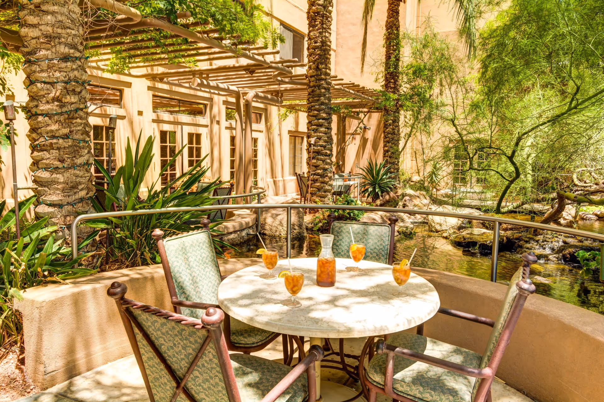 Outdoor courtyard patio with a round table, four chairs and a pitcher of iced drinks beside a pond and palm trees.