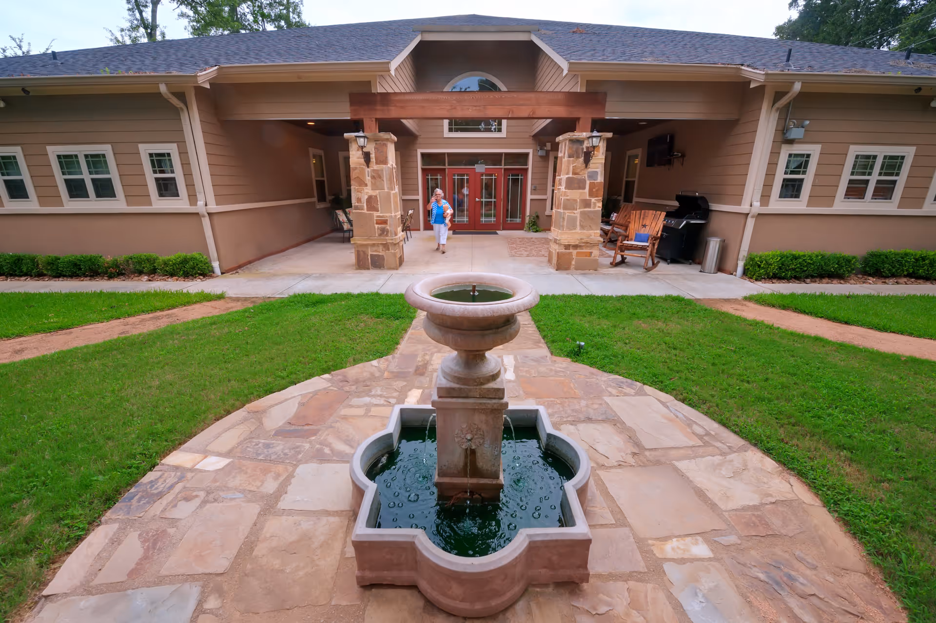 Outdoor courtyard area of Village Green Memory Care Community Conroe featuring a stone water fountain in the center, surrounded by a stone pathway and green grass. The building entrance with double red doors and stone pillars is visible in the background, along with a person walking near the entrance and outdoor seating including a wooden rocking chair and a grill.