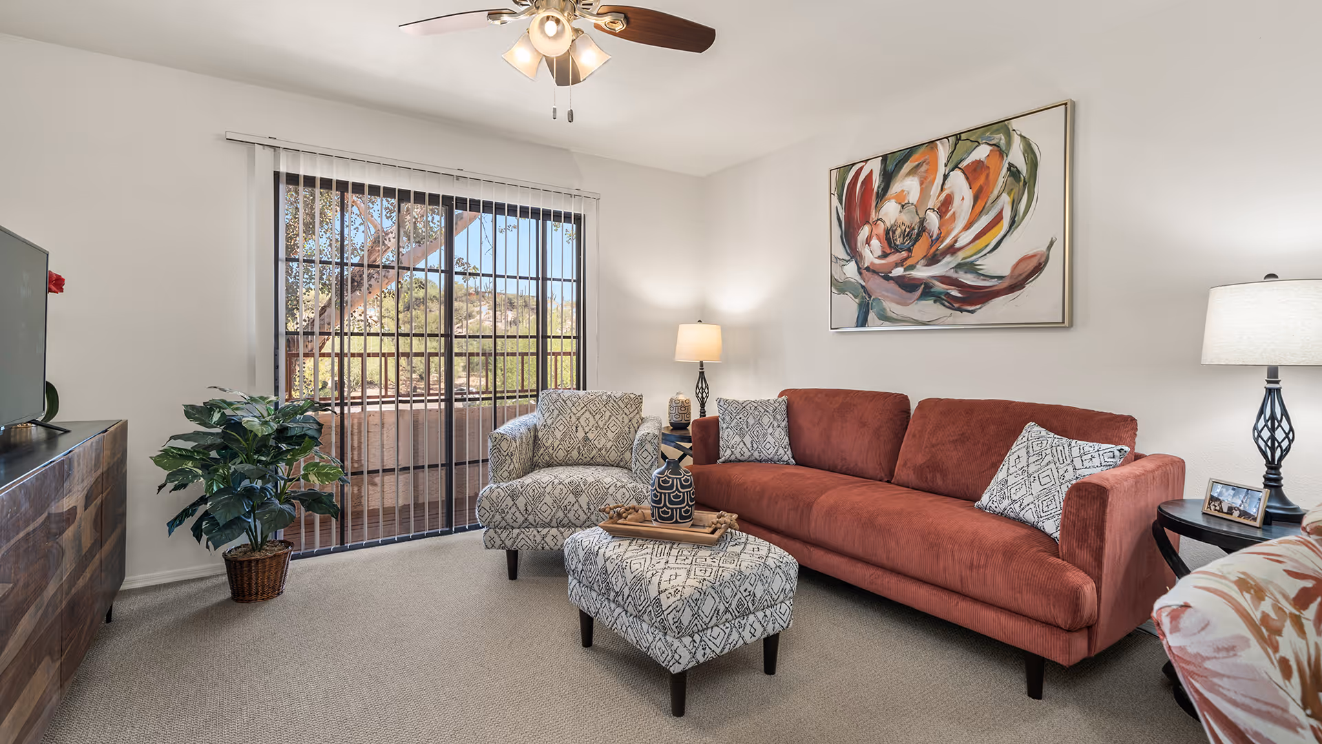 A cozy living room with a rust-colored sofa, a patterned armchair, and a matching ottoman. The room features a ceiling fan with lights, a large window with vertical blinds showing an outdoor view, a wooden TV stand with a television, a potted plant, two table lamps on side tables, and a colorful abstract flower painting on the wall.
