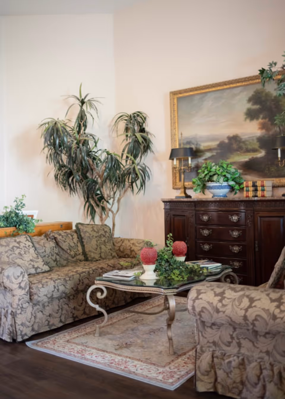 Elegant living room with patterned sofas, a glass-top coffee table, potted plants, and a framed landscape painting above a wooden sideboard.