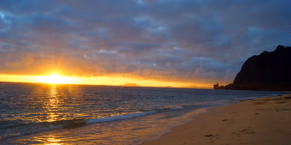 A scenic view of a beach at sunset with the sun partially hidden behind clouds, casting a golden reflection on the ocean waves. A mountain silhouette is visible on the right side of the image.