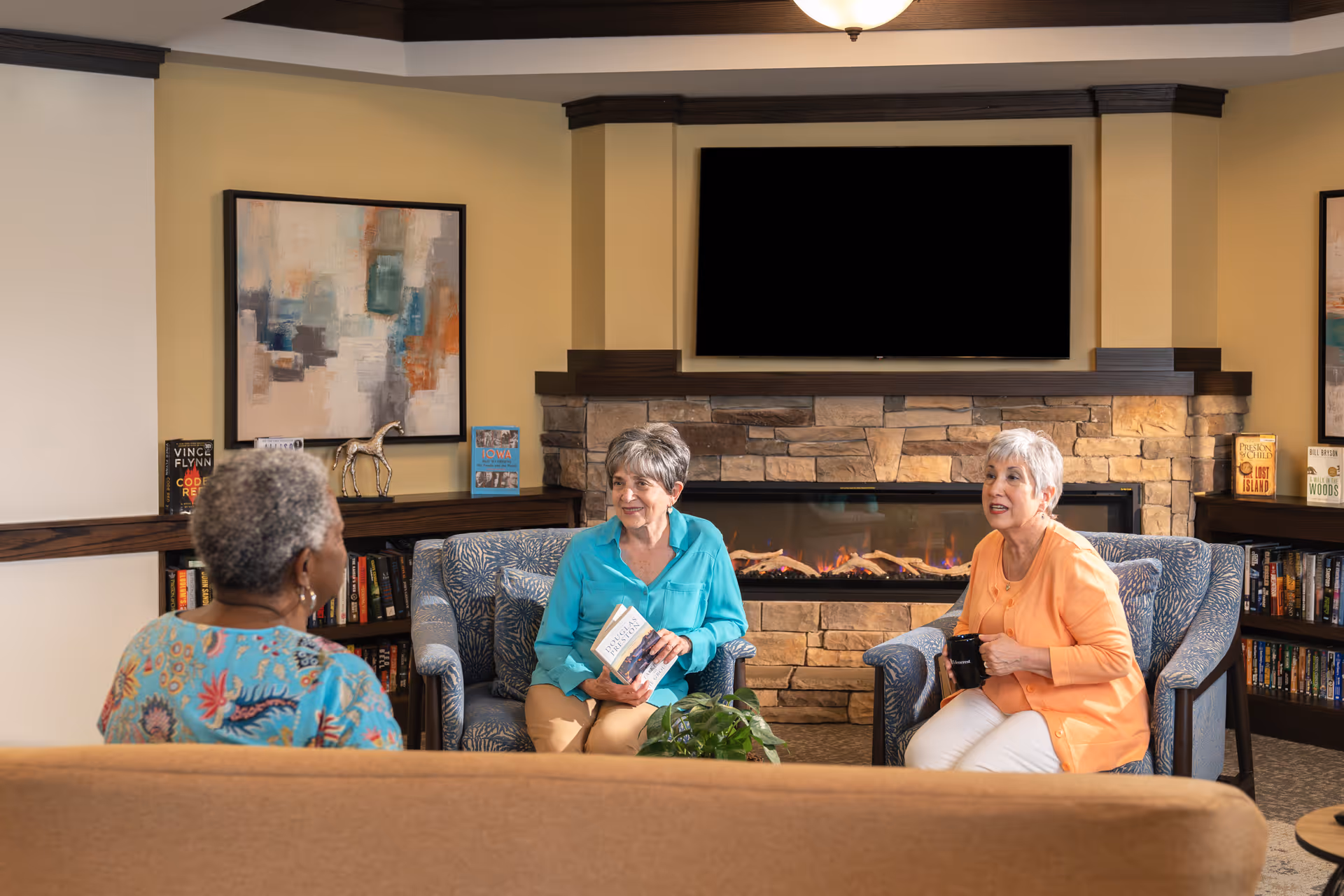 Three elderly women sitting and chatting in a cozy living room with a stone fireplace and a large flat-screen TV mounted above it. Two women are seated in patterned armchairs holding a book and a mug, while the third woman is sitting on a beige sofa with her back to the camera. Bookshelves with books and decorative items flank the fireplace.