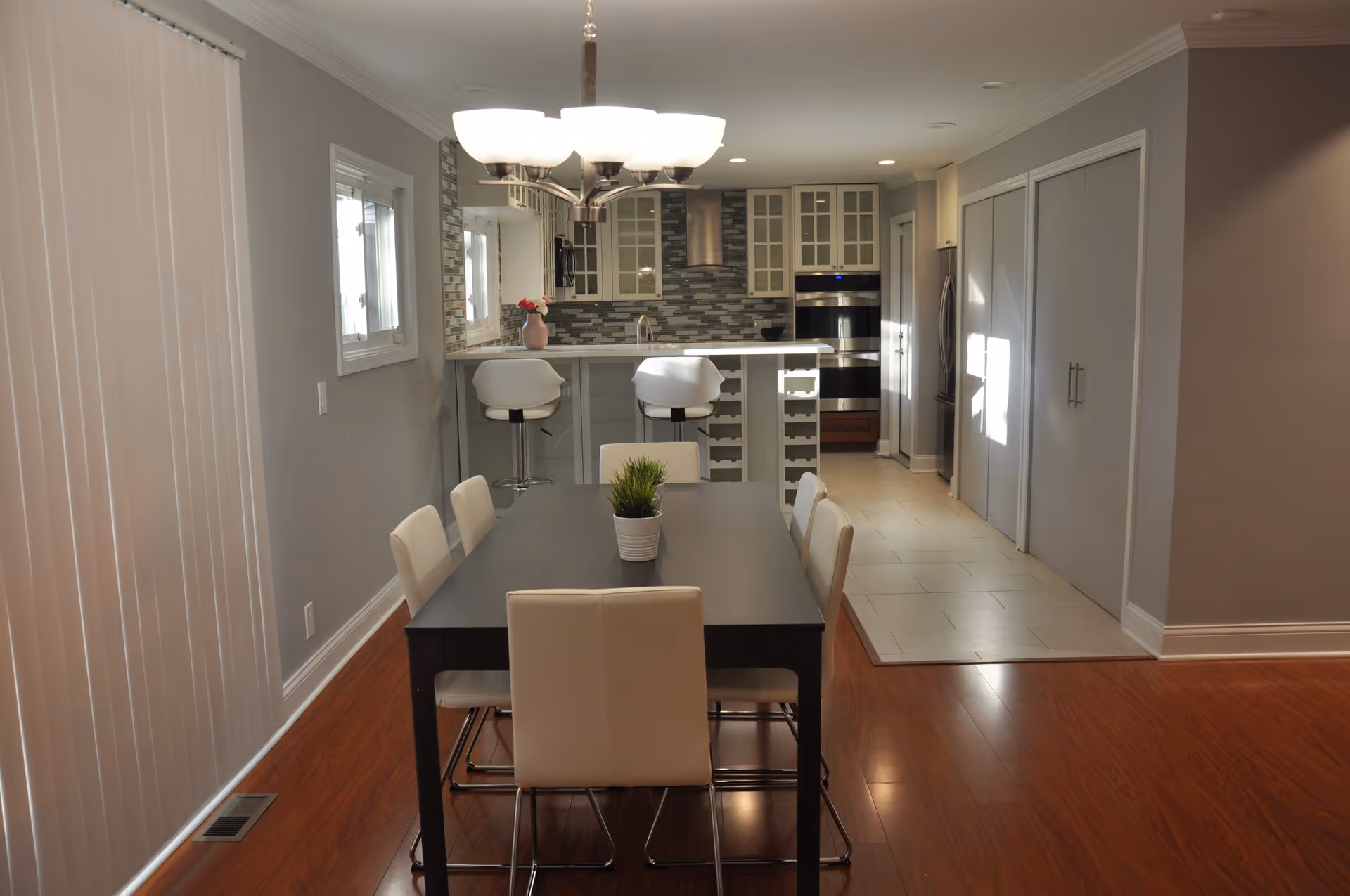 A modern dining area with a dark rectangular table surrounded by six white chairs. A small potted plant is centered on the table. Behind the dining area is a kitchen with a white countertop, two white bar stools, and a backsplash with gray tiles. The kitchen has stainless steel appliances and glass-front cabinets. The floor is a combination of wood and tile, and the walls are painted light gray.