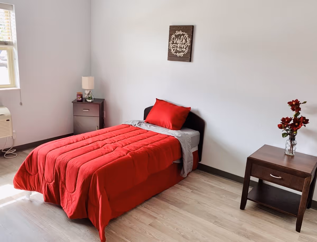 A simple bedroom with a single bed covered in a red comforter and a matching red pillow. There is a small dark wooden nightstand with a lamp and a candle on the left side of the bed, and another small dark wooden table with a vase of red flowers on the right side. A window with blinds is on the left wall, and a decorative wooden sign with the words 'Walk by Faith' hangs above the bed on the white wall.