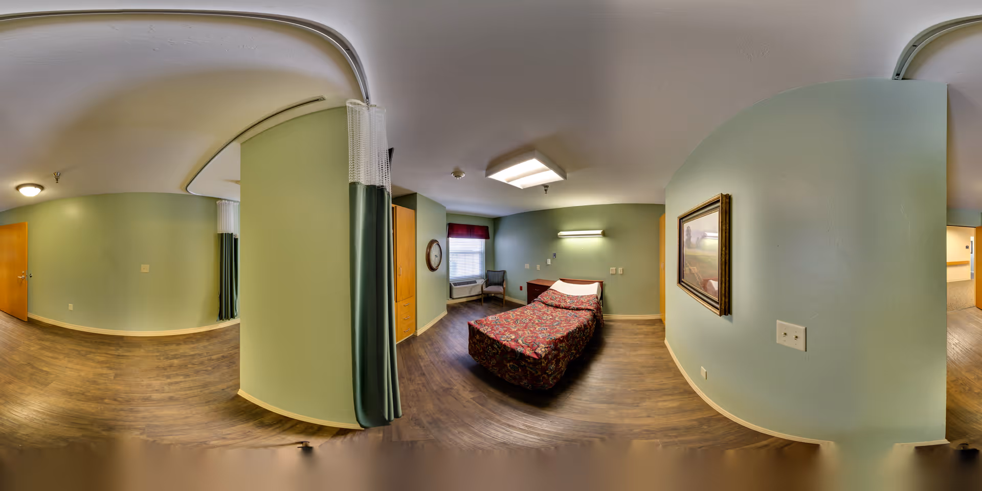 A nursing center bedroom with a single bed covered in a floral bedspread, a chair near a window with a red valance, a wooden cabinet, a wall-mounted light above the bed, and a framed picture on a curved green wall. The room has wood flooring and a privacy curtain on a ceiling track.