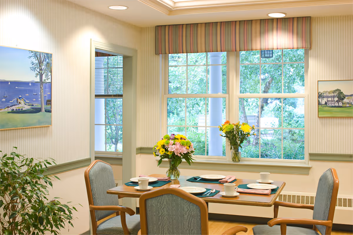 A cozy dining room with a wooden table set for four, including plates, cups, and napkins. Two vases with colorful flowers are placed on the table and windowsill. The room has large windows with a view of green trees outside, light-colored walls with striped wallpaper, and framed paintings on the walls.