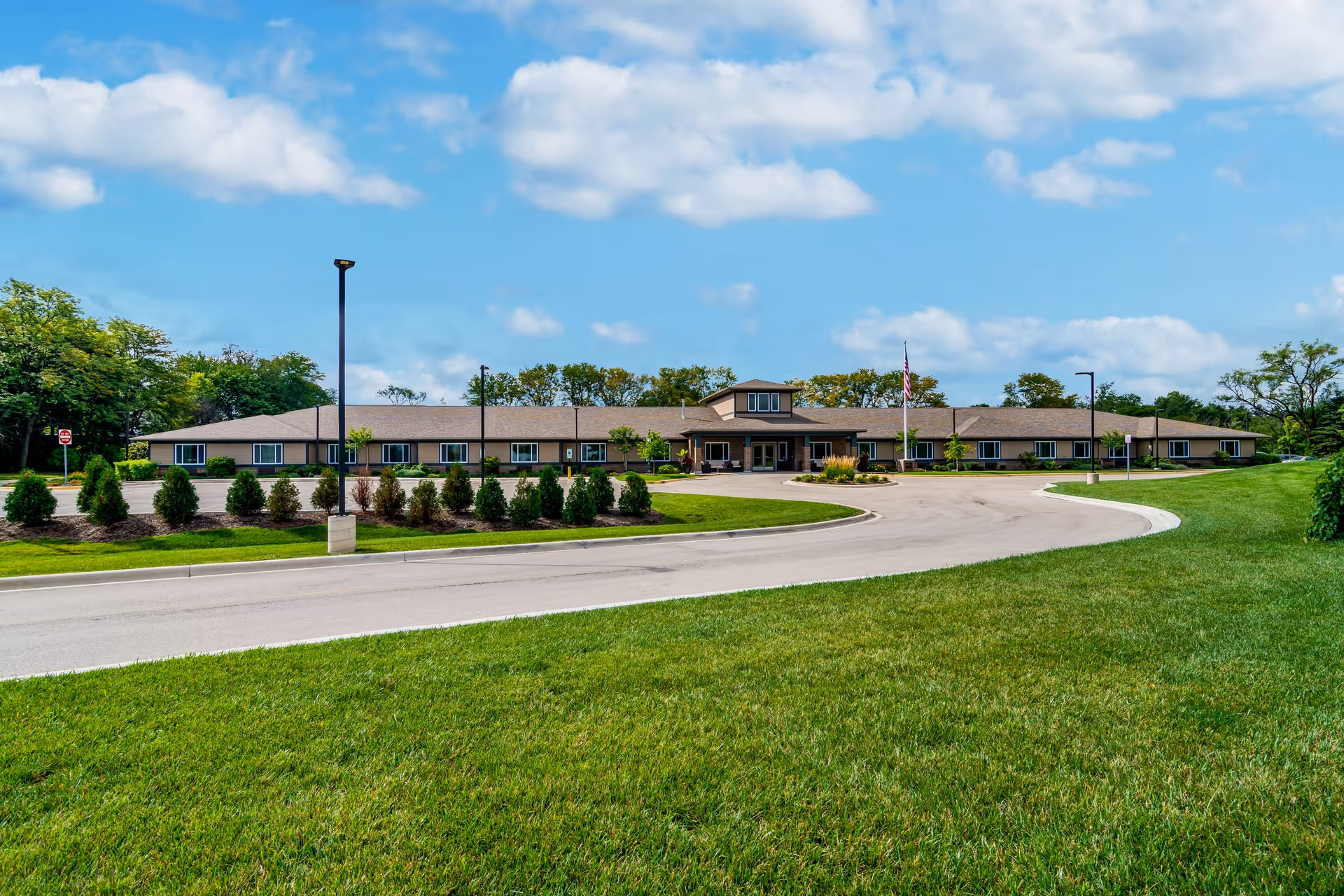 Wide exterior view of a single-story senior living facility building named Harvester Place, surrounded by green grass, small trees, and shrubs under a partly cloudy blue sky. A curved driveway leads to the entrance with an American flag displayed near the front.