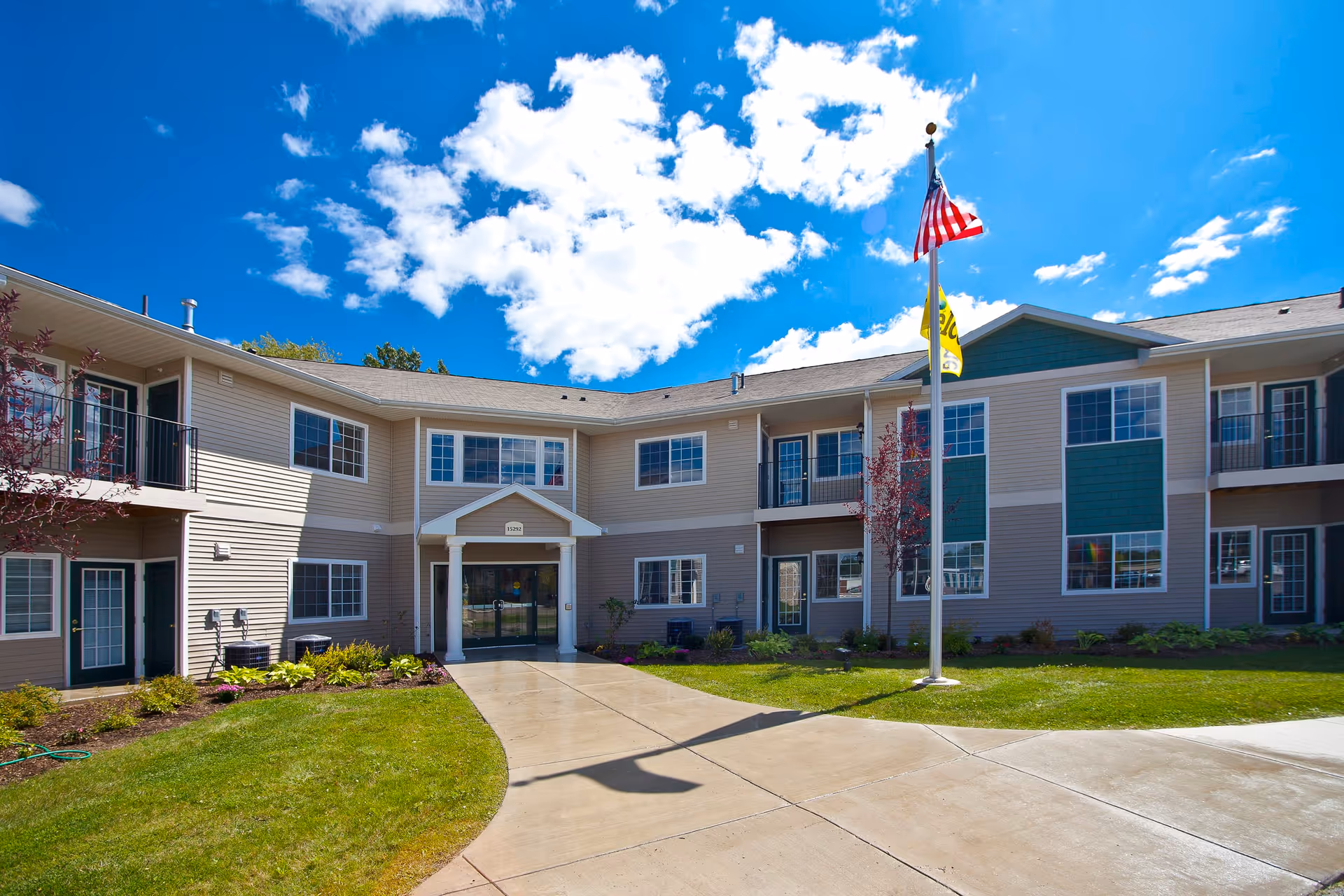 Exterior view of Hawk's Ridge Apartments building under a bright blue sky with scattered clouds. The two-story building has beige and green siding, multiple windows, and balconies. A concrete pathway leads to the main entrance, and an American flag and another flag are flying on a flagpole in front of the building. There is well-maintained grass and landscaping around the pathway.
