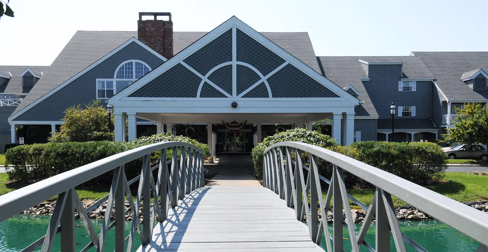 A gray building with white trim and a covered entrance is seen from the perspective of a pedestrian bridge over a small pond. The building has multiple windows and a chimney, with greenery and bushes around the entrance area.