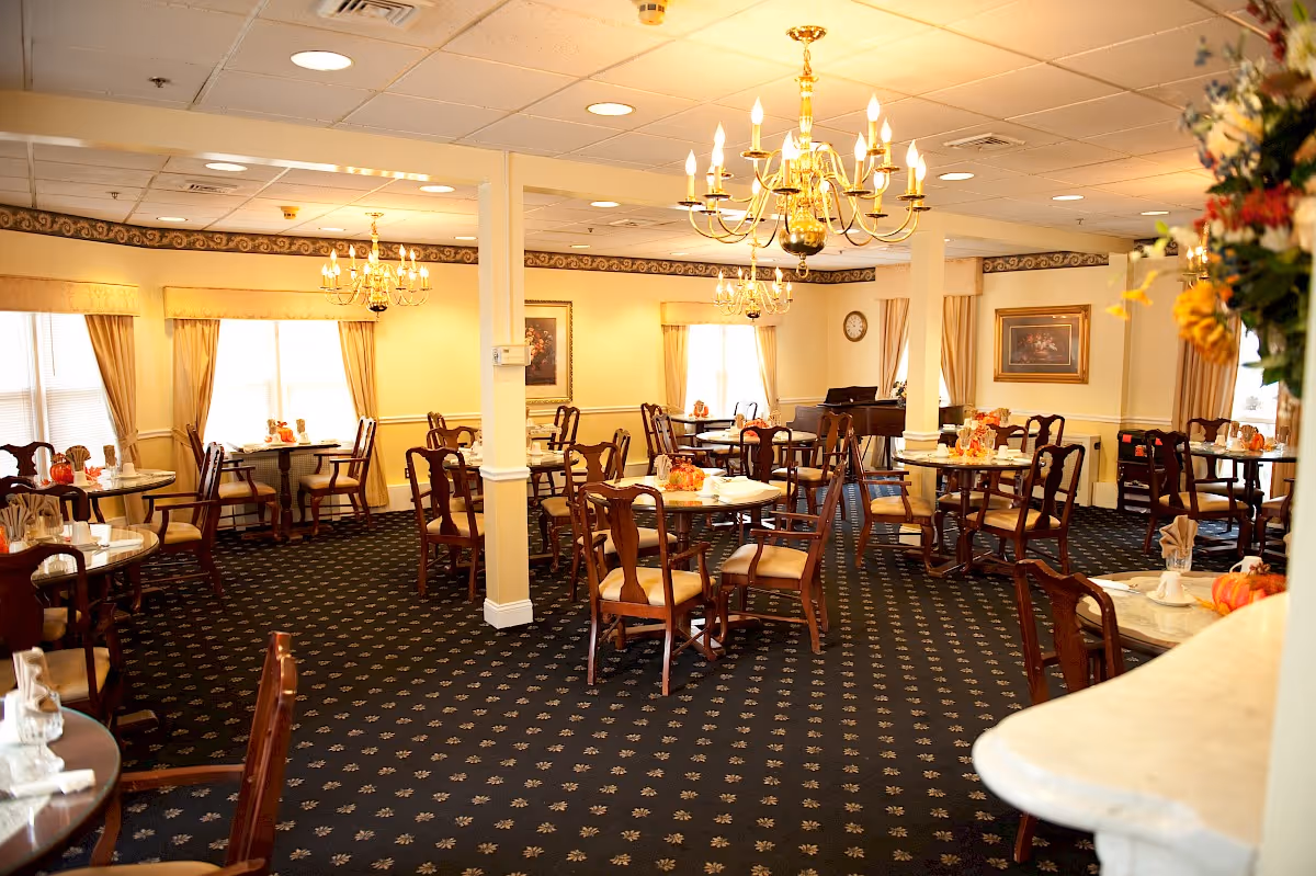 A well-lit dining room with multiple round tables and wooden chairs arranged neatly. The tables are set with napkins and small decorative pumpkins. The room features chandeliers hanging from the ceiling, large windows with beige curtains, framed artwork on the walls, and a piano in the corner.