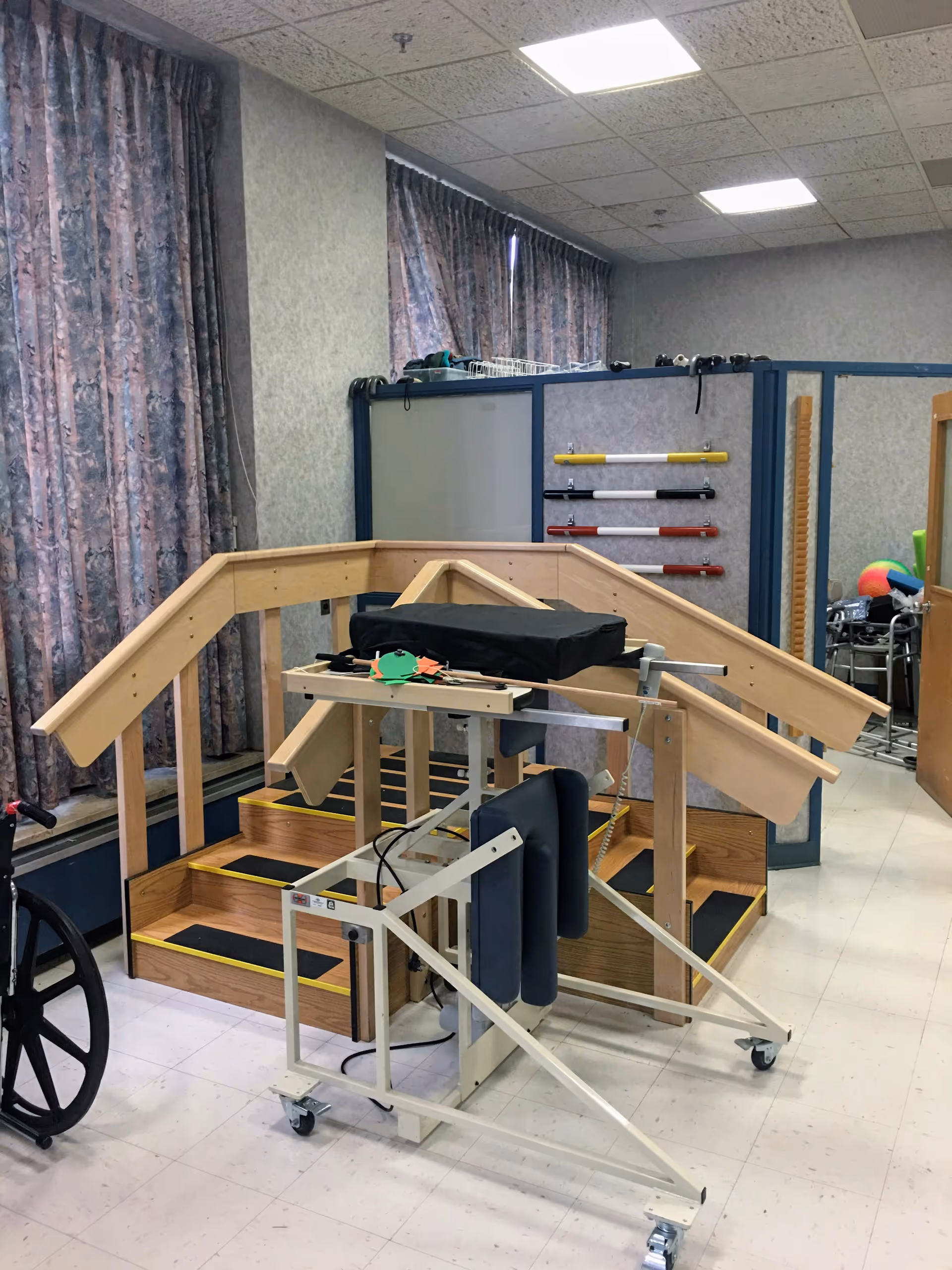Indoor therapy room with wooden practice stairs, parallel rails, and various mobility and rehab equipment.