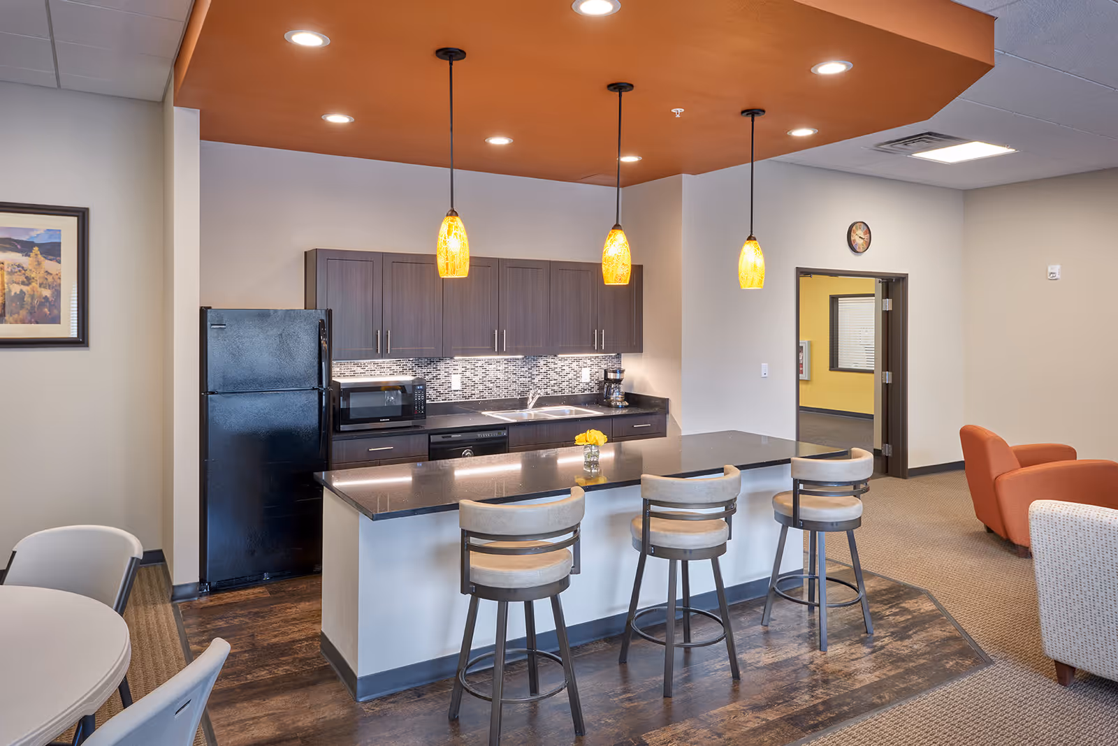A modern kitchen area with a black refrigerator, microwave, coffee maker, and sink. There is a black countertop island with three beige cushioned bar stools. Three pendant lights hang from an orange ceiling section above the island. To the left, there is a round table with chairs, and to the right, a seating area with an orange armchair and a beige patterned chair. A framed picture hangs on the wall, and a clock is above a doorway leading to another room.