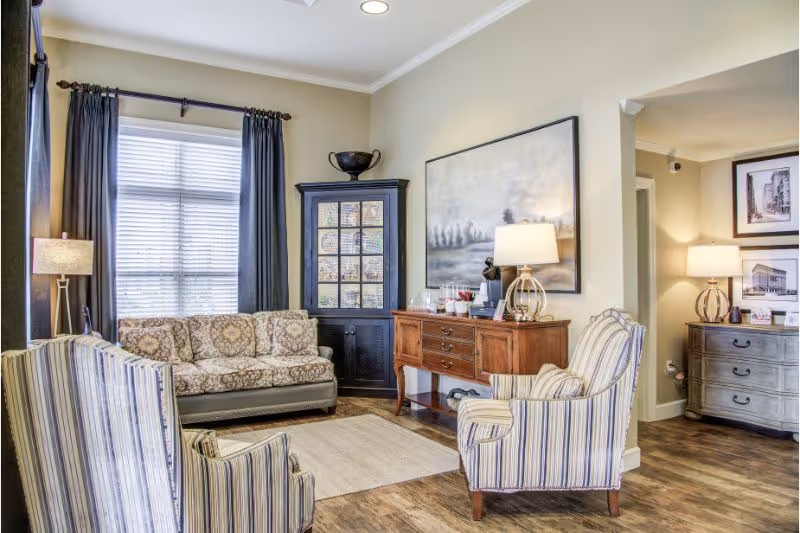 A cozy living room with a patterned sofa, two striped armchairs, a wooden sideboard with a lamp and decorative items, a black corner cabinet, and framed artwork on the walls. The room has large windows with dark curtains and wood flooring.