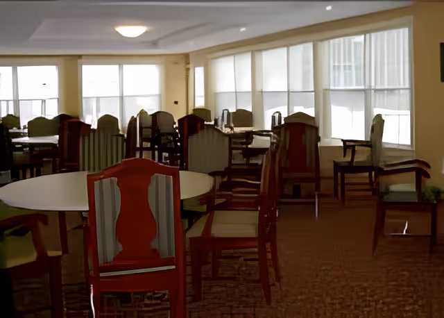 A dining room with multiple round and rectangular tables surrounded by wooden chairs with green cushions. Large windows with white blinds allow natural light to fill the room.