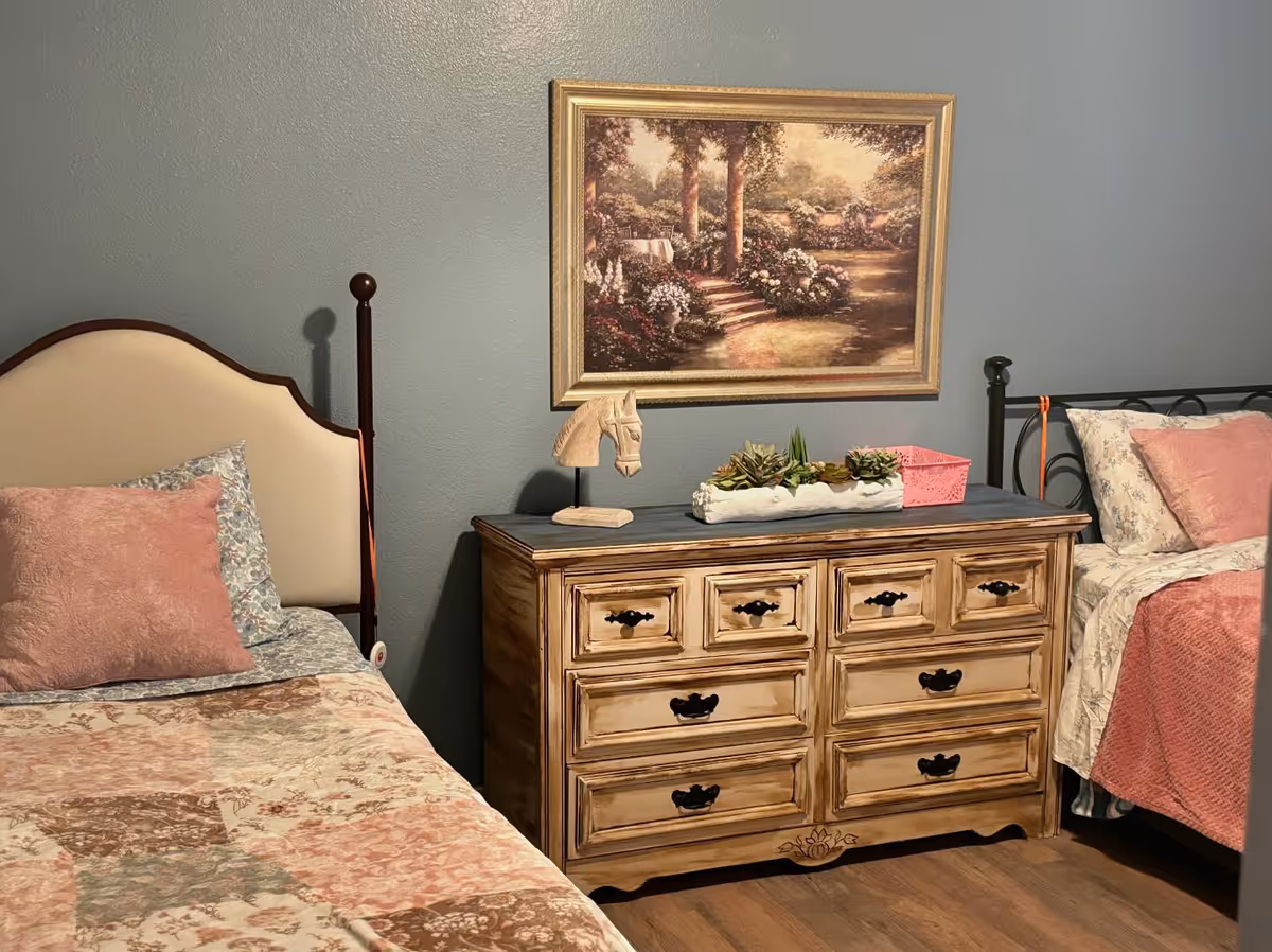 A cozy bedroom with two twin beds on either side of a wooden dresser. Each bed has floral-patterned bedding with pink pillows. On the dresser, there is a decorative horse head sculpture, a planter with succulents, and a pink basket. Above the dresser hangs a framed painting of a garden scene with trees, flowers, and a pathway.