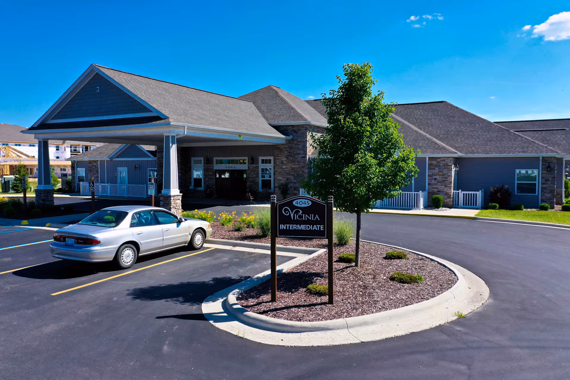 Exterior view of Vicinia Gardens Luxury Retirement Living - Intermediate Care building with a covered entrance, a parked silver car, a small landscaped island with a tree and bushes, and a clear blue sky.