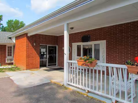 Red brick Elms Health Care Center entrance with white railing, porch seating, and flowers.