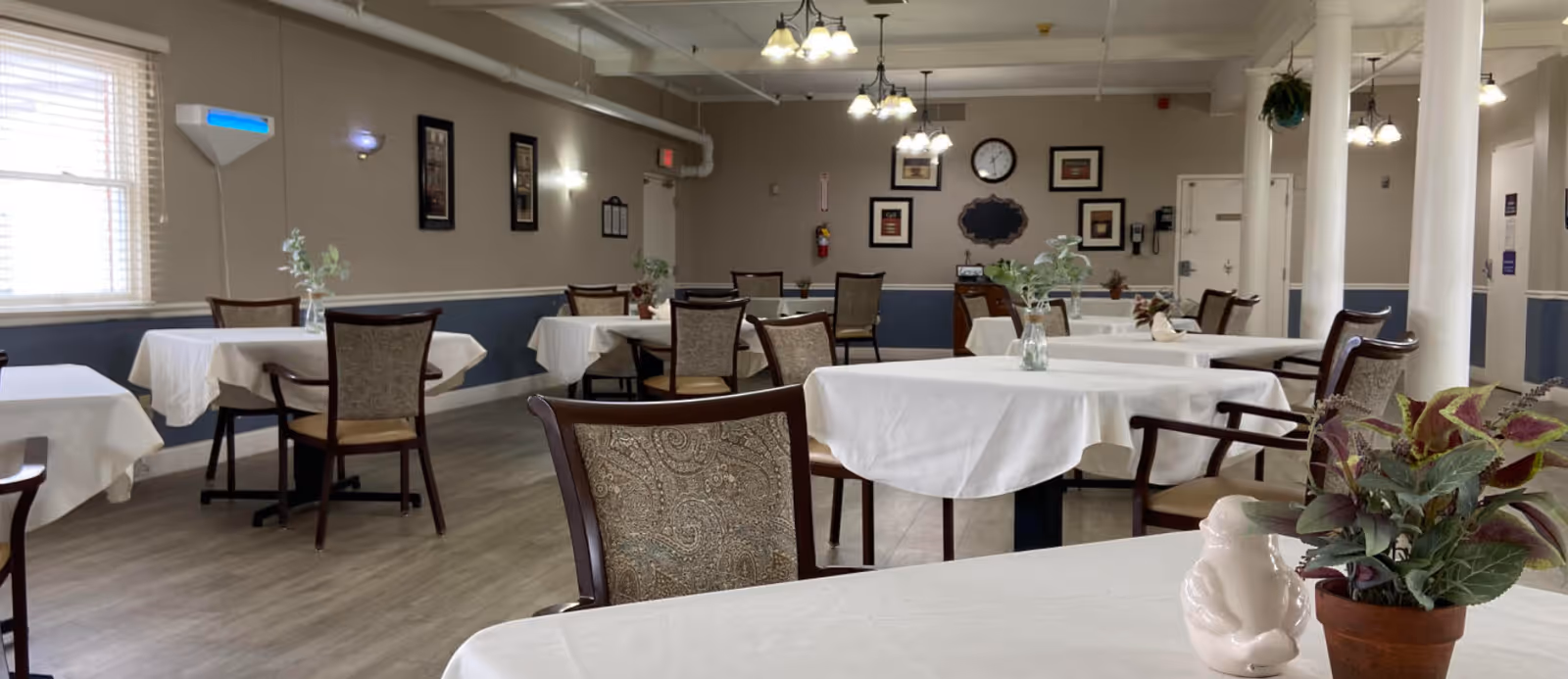 A dining room with several tables covered in white tablecloths, each surrounded by wooden chairs with patterned upholstery. Small plants and decorative items are placed on the tables. The room has light-colored walls with framed pictures and a clock on the far wall. Ceiling lights hang from above, and there are windows letting in natural light on the left side.