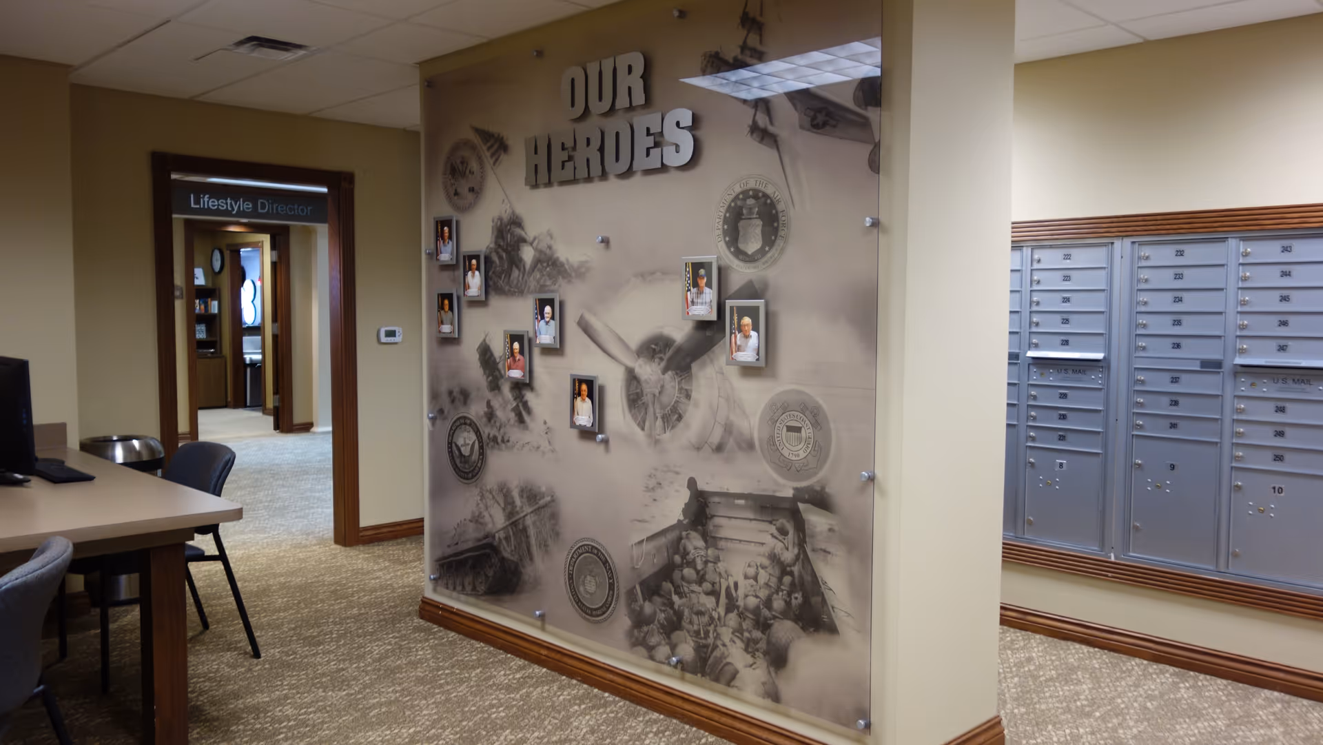 Interior view of a senior living facility hallway with a wall display titled 'OUR HEROES' featuring photos of individuals and military-themed artwork. To the right, there are multiple mailboxes mounted on the wall. On the left, there is a desk with chairs and a doorway labeled 'Lifestyle Director' leading to another room.