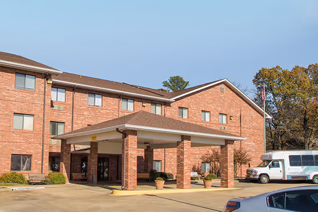 Exterior view of a three-story brick building with a covered entrance supported by brick columns. There are potted plants near the entrance, a flagpole with an American flag, and a white shuttle bus parked to the right. The sky is clear and blue.