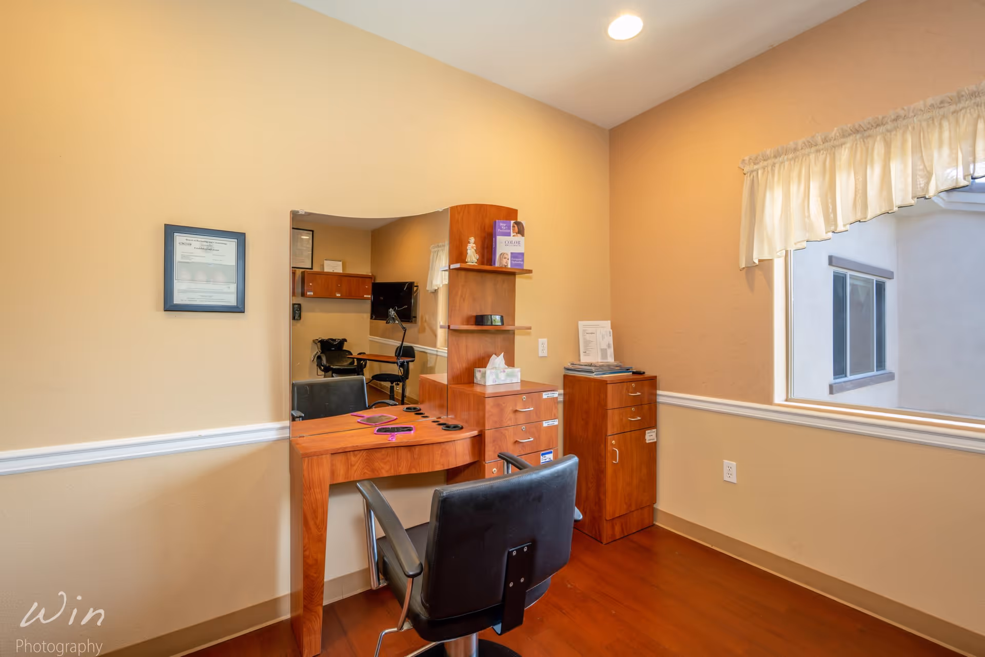 A small room with a wooden vanity table and a black swivel chair in front of a large mirror. The vanity has shelves with brochures and a tissue box. There is a framed certificate on the beige wall and a window with a white curtain letting in natural light. The floor is wooden and there is a small cabinet next to the vanity.