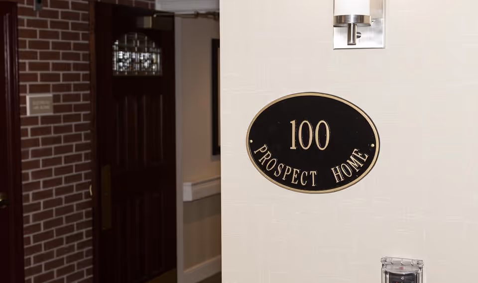 Interior hallway corner with a black oval plaque displaying the number 100 and the words 'Prospect Home' mounted on a light-colored wall. A modern wall sconce light fixture is above the plaque. To the left, there is a dark wooden door with a glass panel and a brick wall section.