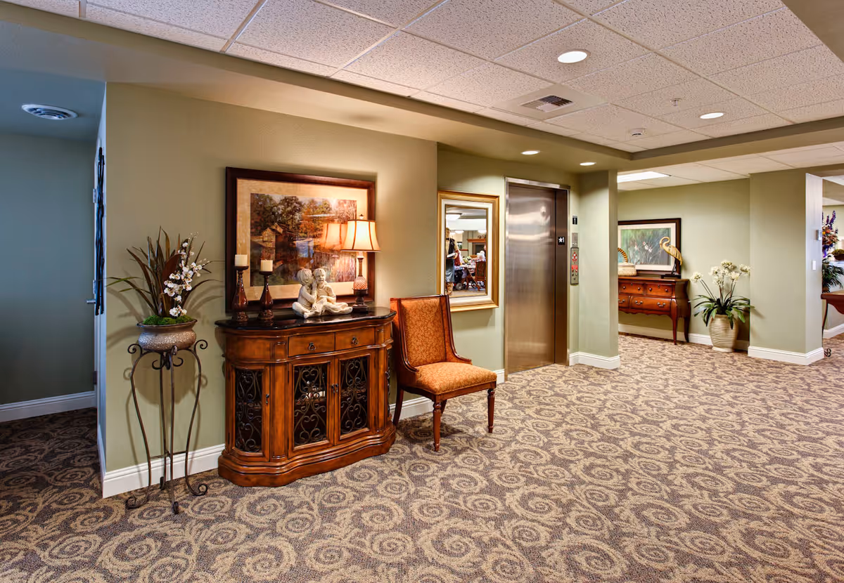 Interior hallway of a senior living community with patterned carpet, green walls, and a ceiling with recessed lighting. There is a wooden cabinet with decorative items including a lamp, candles, and a sculpture of two children reading. Next to the cabinet is an upholstered chair. An elevator with stainless steel doors is visible, along with framed artwork and potted plants placed around the hallway.