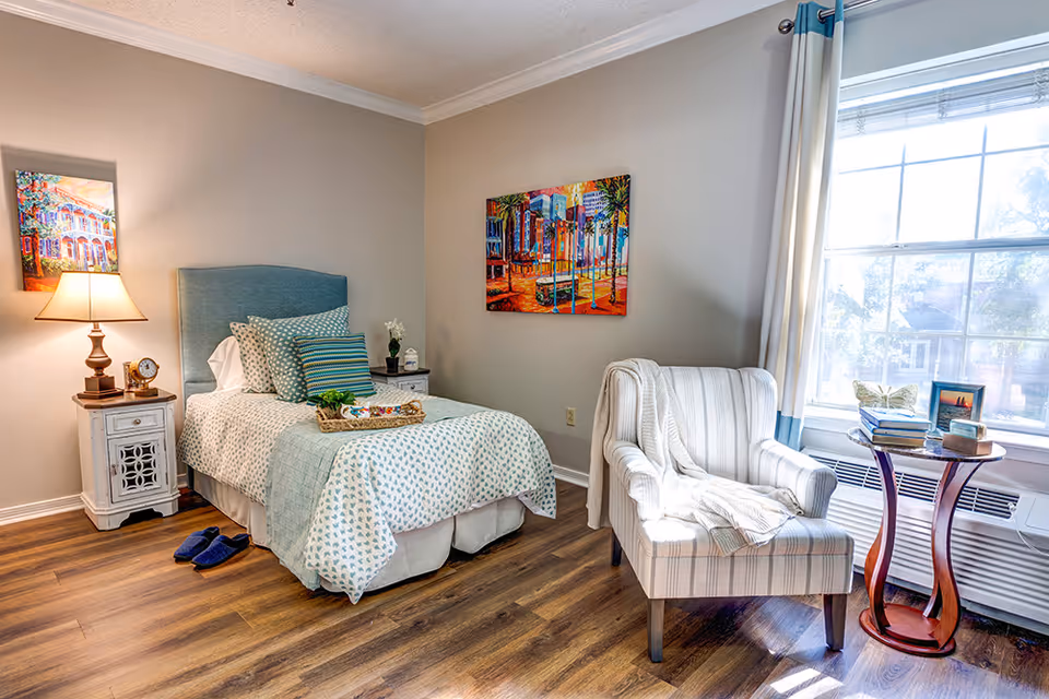 A cozy bedroom with a single bed featuring a blue upholstered headboard, patterned bedding, and multiple pillows. Next to the bed is a white nightstand with a lamp, clock, and decorative items. A striped armchair with a white throw blanket is positioned near a large window with white and blue curtains. A small round wooden table beside the chair holds books and framed pictures. The room has wooden flooring and colorful artwork on the walls.