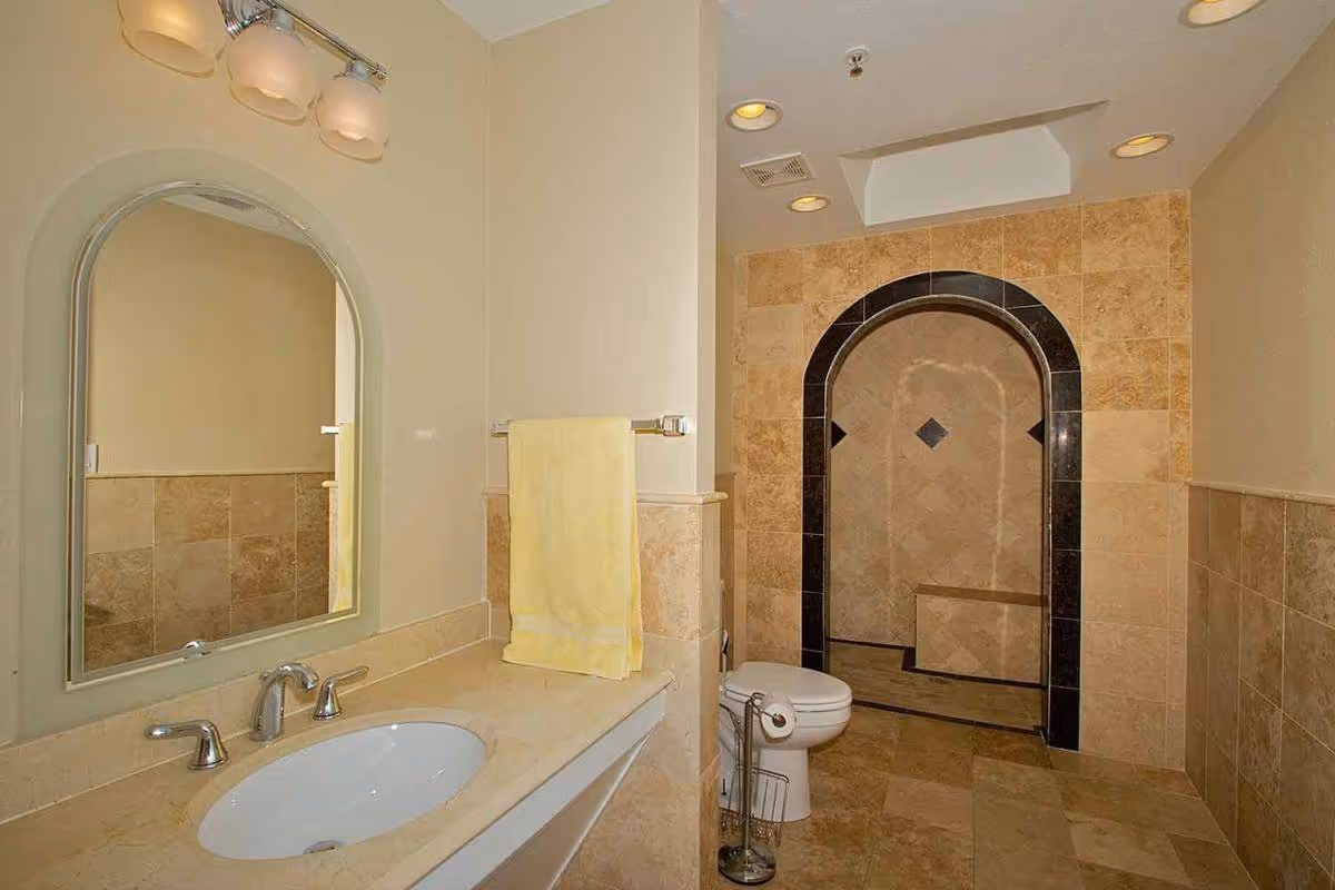 A bathroom with beige tiled walls and floor, featuring a white sink with a silver faucet and a large arched mirror above it. A yellow towel hangs on a rack next to the sink. There is a white toilet next to an arched walk-in shower with decorative black and beige tiles.