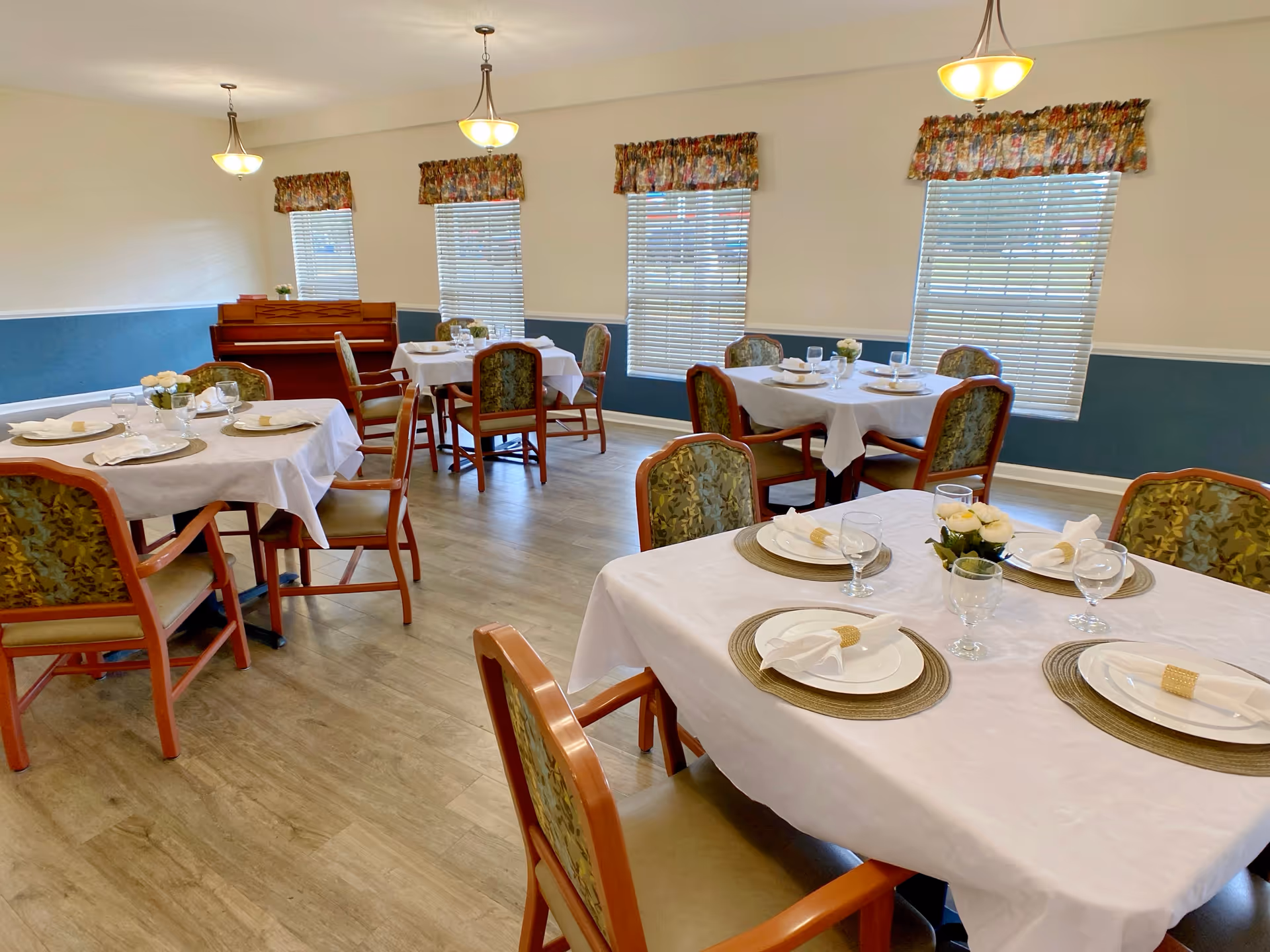 A bright dining room with several round and square tables covered with white tablecloths. Each table is set with plates, napkins, glasses, and small flower arrangements. The room has wooden chairs with patterned upholstery, three windows with floral valances, and a piano against the far wall. The floor is light wood, and the walls are painted white with a blue wainscoting.