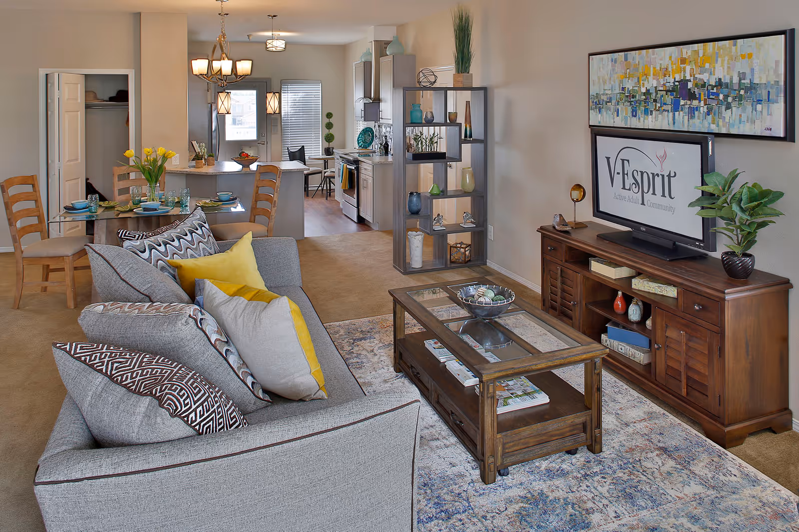 A cozy living room and dining area in an apartment at V-Esprit. The living room features a gray sofa with patterned and yellow pillows, a wooden coffee table with decorative items, and a wooden TV stand with a flat-screen TV displaying the V-Esprit logo. There is a colorful abstract painting above the TV. The dining area has a glass-top table set with dishes and glasses, surrounded by wooden chairs. The kitchen is visible in the background with white cabinets and stainless steel appliances. The space is decorated with plants and modern light fixtures.