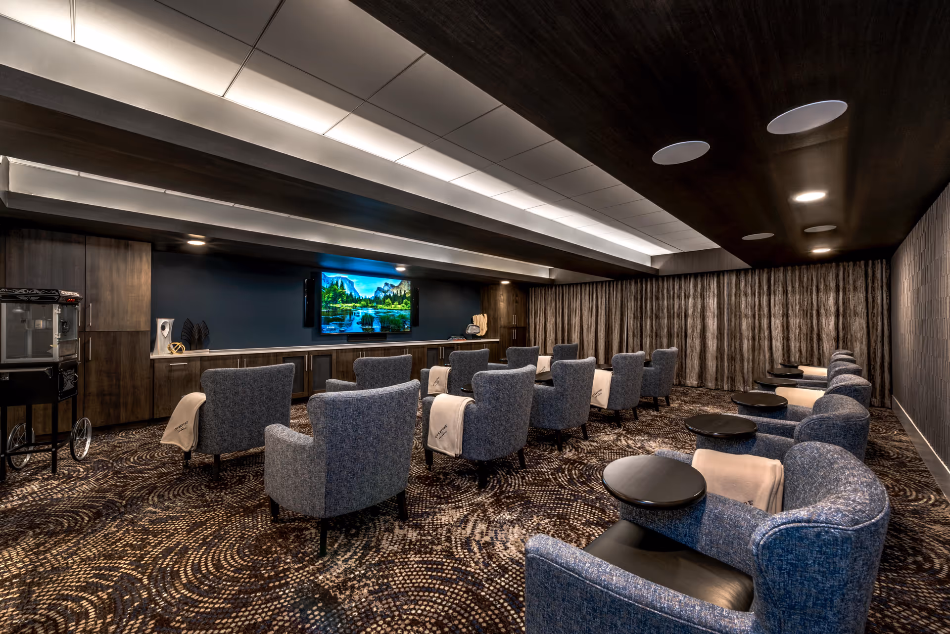 A cozy theater room with multiple rows of blue upholstered armchairs, each equipped with a small black side table and a beige blanket draped over the back. The room features a large flat-screen TV mounted on a dark blue wall with wooden cabinets below. The ceiling has recessed lighting and acoustic panels, and the floor is covered with a patterned carpet. A popcorn machine is visible on the left side of the room.