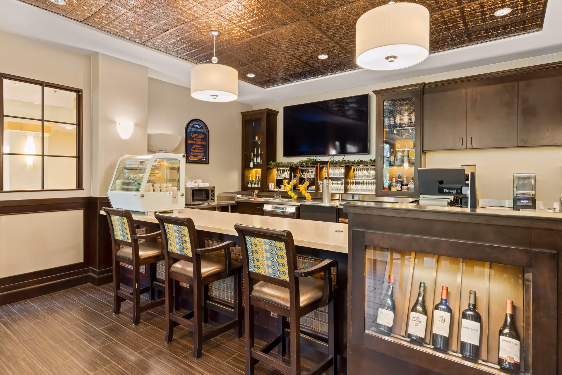 Interior dining/bar area with a long counter, four barstools, a display case, wine bottles and a wall-mounted TV.