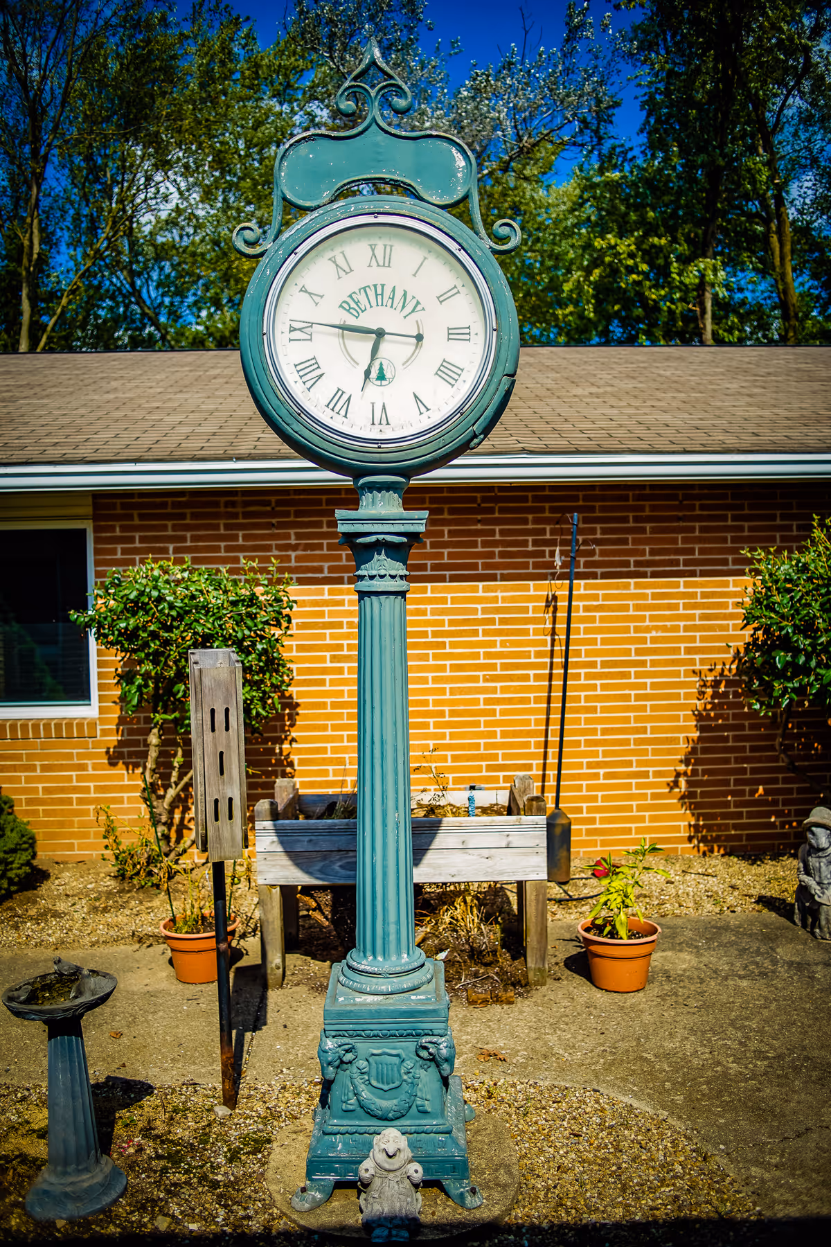 An outdoor vintage-style clock on a tall green post with the word 'BETHANY' on its face, standing in front of a brick building with a window and surrounded by potted plants and garden decorations under a clear blue sky.