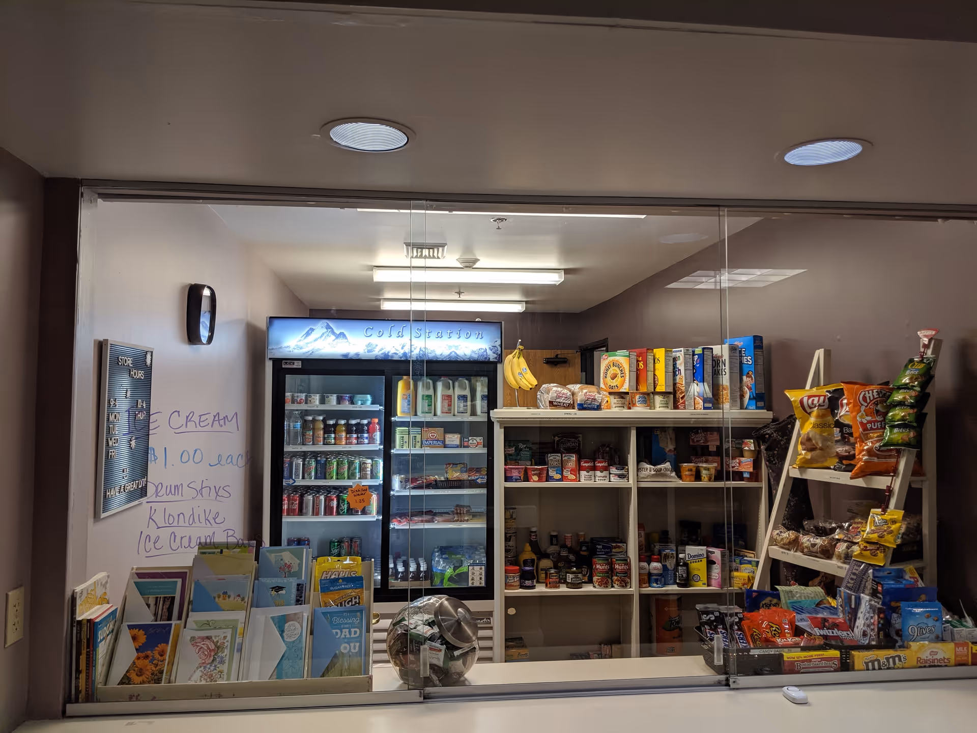 A small convenience store area inside a facility with shelves stocked with various snacks, canned goods, and groceries. There is a refrigerated display labeled 'Cold Station' containing drinks and dairy products. A handwritten sign on the wall advertises ice cream for $1.00 each. Greeting cards are displayed on the left side, and a glass partition separates the store area from the viewer.