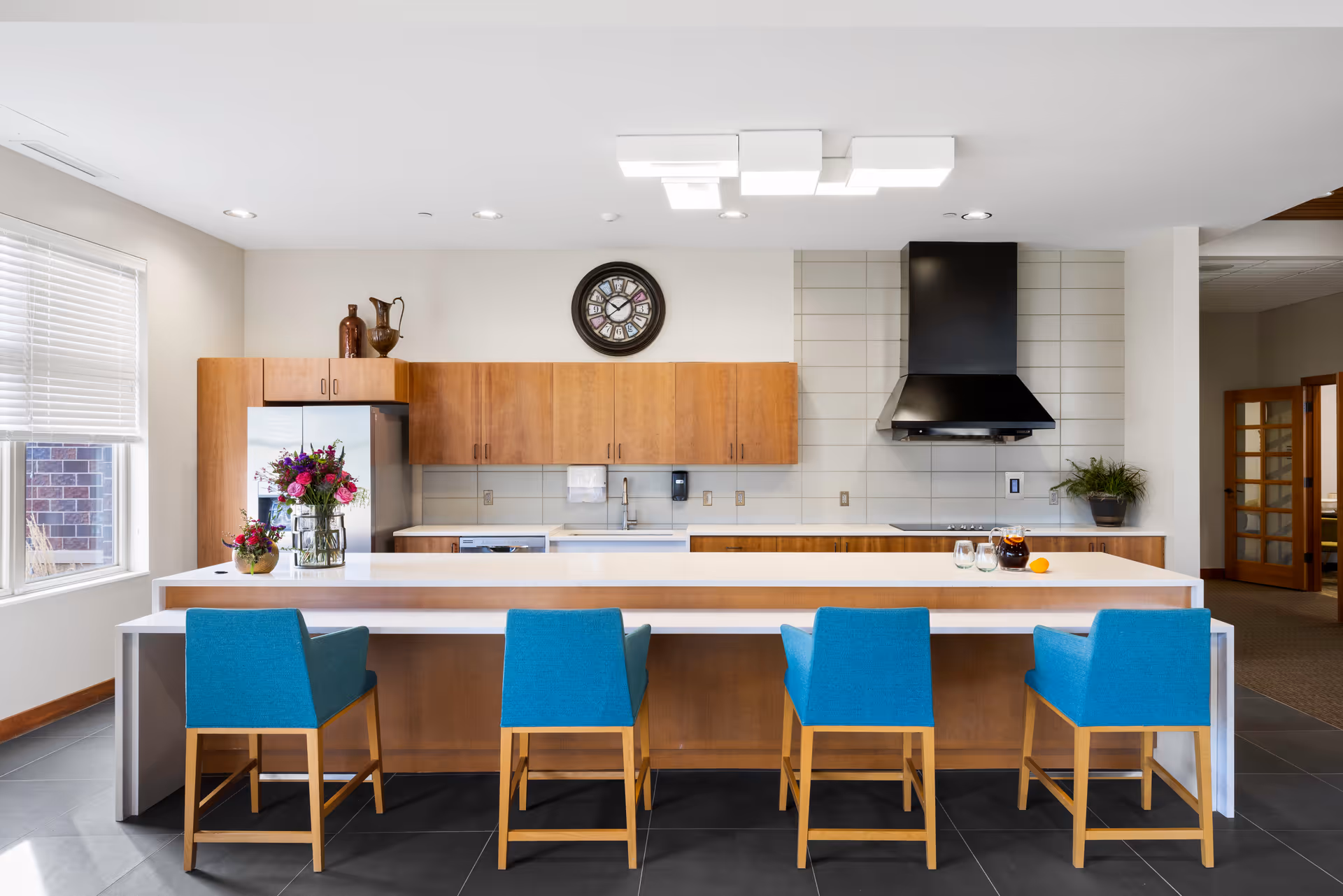 Modern kitchen area with a long white island countertop and four blue chairs with wooden legs. The back wall has wooden cabinets, a stainless steel refrigerator, a sink, and a black range hood above a stovetop. A round clock is mounted on the wall above the cabinets. There are decorative items including flowers in vases on the countertop and cabinets. A window with blinds is on the left side, and an open doorway is visible on the right.