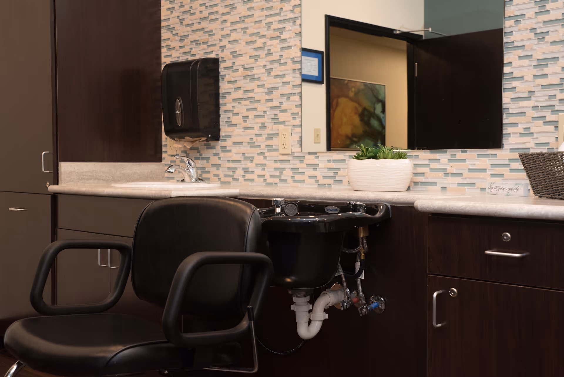 Interior view of a salon or grooming station with a black salon chair in front of a black wash basin. The countertop has a white sink, a small potted plant, and a basket. The wall behind features a mosaic tile backsplash and a large mirror reflecting part of the room.