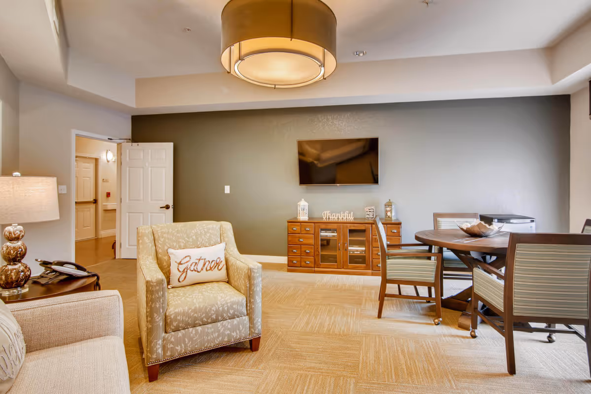 A cozy living room area with a beige armchair featuring a 'Gather' pillow, a beige sofa, a wooden side table with a lamp and phone, a wooden cabinet with decorative items and a mounted flat-screen TV on a gray accent wall, and a round wooden dining table with four chairs on a carpeted floor.