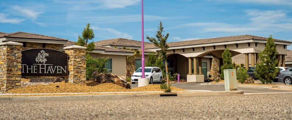 Exterior view of The Haven at Sky Mountain Independent and Assisted Living Community showing the main entrance with stone pillars and a sign displaying the facility name and address. Several cars are parked near the entrance under a clear blue sky.