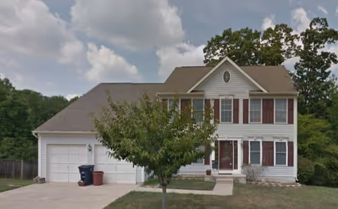 Two-story white house with brown shutters and a brown roof, a small tree in the front yard, and a driveway leading to a double garage. The house is surrounded by greenery and trees in the background under a partly cloudy sky.