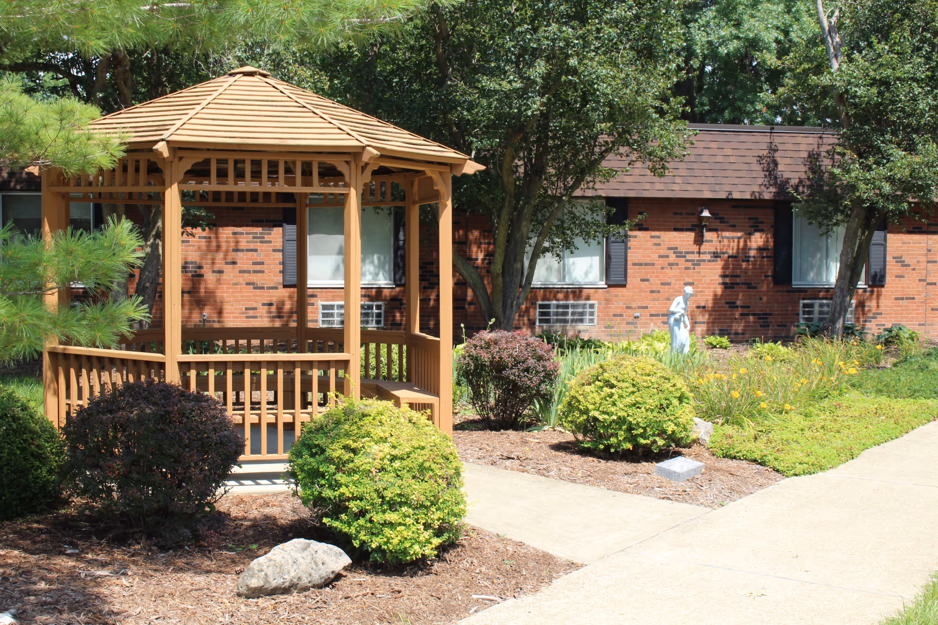 A wooden gazebo surrounded by bushes and trees in a garden area outside a brick building with windows. A concrete pathway runs alongside the garden, and a small statue is visible among the plants.