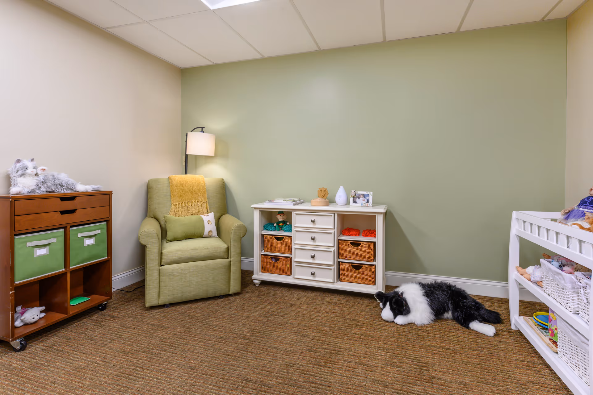 A cozy room with a green armchair draped with a yellow throw blanket, a wooden storage unit with green fabric bins, a white cabinet with wicker baskets, and a white shelving unit holding toys and stuffed animals. A black and white stuffed dog lies on the carpeted floor. The walls are painted light green and beige.